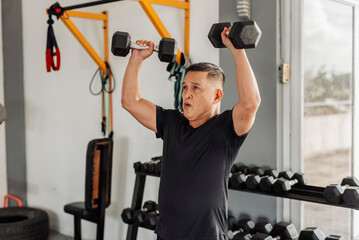Southeast Asian man in his mid 50s performs an overhead dumbbell shoulder press at an open-air gym with proper form and dedication.