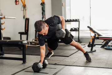 Southeast Asian man in his 50s performs renegade rows on the gym floor, training core and upper body strength with full effort.