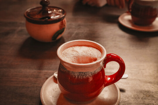 A cup of coffee in a rustic red ceramic mug on a wooden table setting.