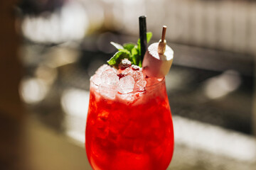 Refreshing red cocktail drink. Glass with ice and garnish on blurred bar background. Summer beverage with mint and fruit skewer. Tasty alcohol refreshment. Evening drink in warm light setting.