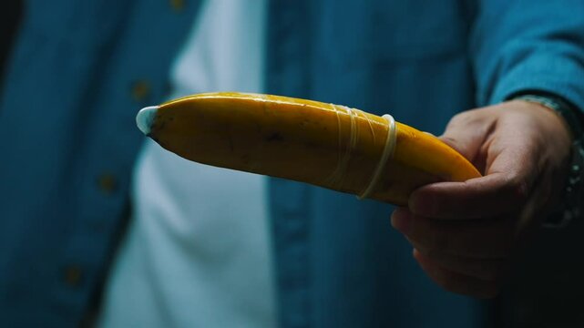 A man in a dark room with artificial lighting holds a ripe banana in his hands with a condom on.
Condom on a banana.
Health, sexually transmitted diseases, sex education
