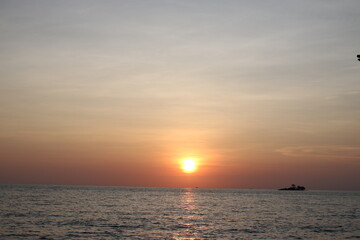 Sunset over a calm ocean with soft clouds and warm tones. A small island silhouettes the horizon, creating a tranquil and scenic seascape perfect for travel, nature, or relaxation themes.

