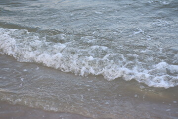 wave rolls onto the sandy shore, with soft white sea foam forming at the edge. The water is a calm blue-gray, reflecting the sky, capturing a peaceful coastal moment.