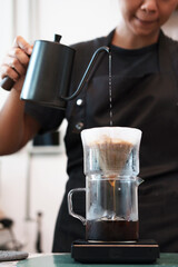 Close up Professional Asian woman barista wearing black apron pouring hot water into a coffee pitcher on counter of cafe
