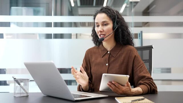 Female corporate operator in headset working in customer support service on helpline telesale talking using laptop and tablet in business office. Agent of call center advises client, answer questions