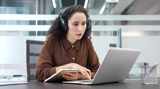 Young businesswoman in wireless headphones watching video call online conference using laptop taking notes sitting in office. Female employee listening to remote business training, course or seminar