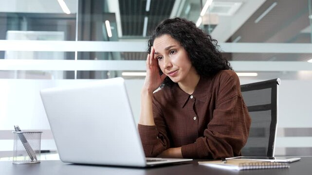 Upset shocked businesswoman reads bad news on a laptop while sitting at a desk at workplace in a modern business office. Frustrated worried woman is disappointed by a negative message on the computer