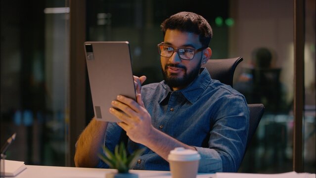 Indian business man talking gadget digital tablet online conference video call chatting with device in evening night office male businessman ethnic guy executive manager conversation virtual meeting