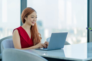 Smiling Asian businesswoman in red dress working remotely with laptop in modern office with panoramic city view, showing confidence and positive energy.
