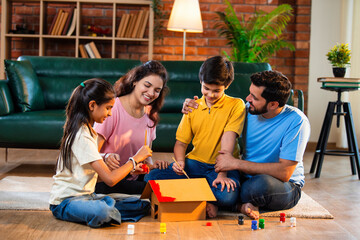Siblings colour cardboard house model on floor while parents watch, bonding as family at home