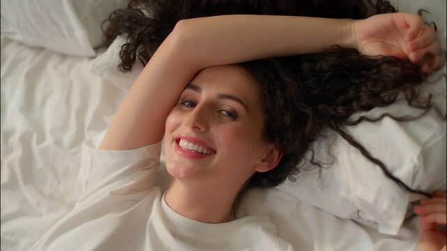 Caucasian happy woman curly girl relaxing in bed cozy home bedroom beautiful female pampering smile awake morning weekend in hotel apartment wake up soft mattress pillow health enjoy rest comfortable