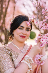 A woman in a shimmering pink traditional outfit stands serenely among cherry blossoms, radiating warmth, elegance, and cultural pride.