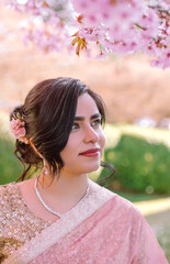A woman in a shimmering pink traditional outfit stands serenely among cherry blossoms, radiating warmth, elegance, and cultural pride.