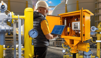 Engineer in hard hat operates a laptop while inspecting an industrial gas control panel, ensuring...