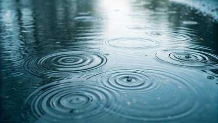 Raindrops create ripples and concentric circles on the surface of a puddle, with a dark, blurred reflection of the surroundings visible, symbolizing rain, weather, water, tranquility