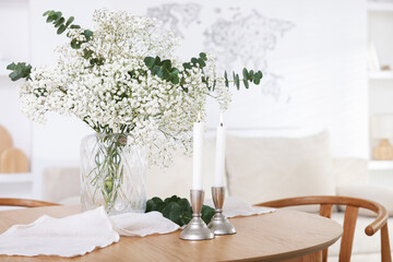 Bouquet of beautiful gypsophila flowers with eucalyptus branches in vase and burning candles on wooden table indoors