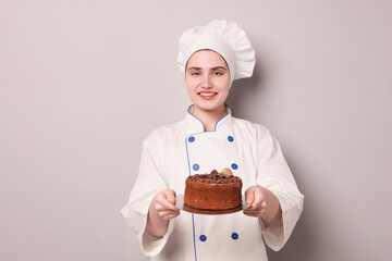 Confectioner in uniform holding delicious chocolate cake on light grey background