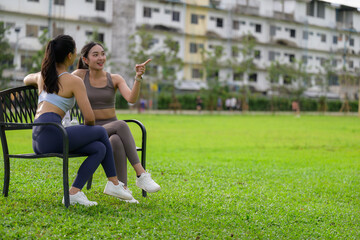 Fototapeta premium Two fit women taking a break on a bench in a green park after training, enjoying conversation and pointing at something