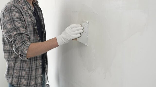 Unknown construction worker man wearing gloves and plaid shirt is using a putty knife to apply plaster on a white wall, during a home renovation or construction project