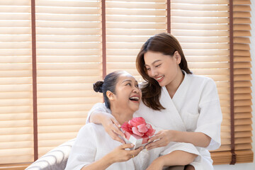 gift, mother, day, bonding, skincare: Laughing Asian daughter hugs delighted mom in white robes, offering pink gift box to celebrate Mother Day and share relaxing spa love, health, glowing joy