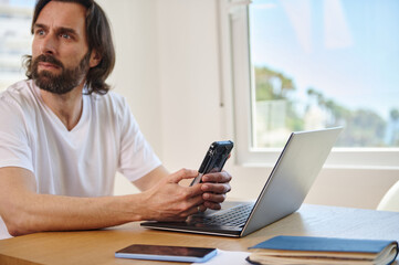 Relaxed Young Man Working Remotely Using Laptop and Smartphone in Minimalist Home Office