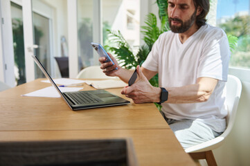 Bearded Man Working Remotely While Using Laptop and Smartphone in Modern Home Office