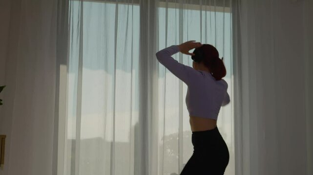 A woman wearing headphones enters her living room with joy, dancing as she gets ready to engage in her morning workout on a fitness mat, promoting a healthy lifestyle.