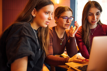 Teenagers are discussing ideas and working on a laptop in a cafe. The girls look at the screen and talk about the educational project. The friends study in the cozy interior.