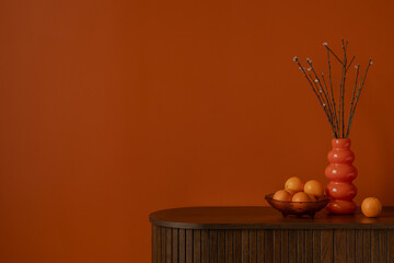 Bold orange wall with wooden sideboard, sculptural vase, and fresh oranges. Minimalist still life with warm tones and natural textures. Copy space.