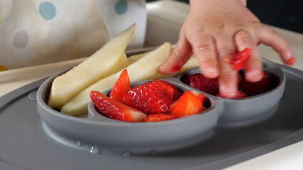 Small dirty hand of a baby trying to eat fresh fruit with his hands, taking fruit from a grey silicone plate