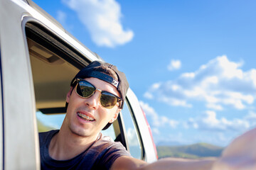 Young Brazilian man taking a selfie from a car window during a road trip. Summer sky, sunglasses, and fun vibes. Concept of freedom, adventure, and youth travel