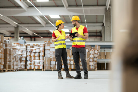 Male and female workers in protective helmets reviewing information on a digital tablet in a large warehouse.