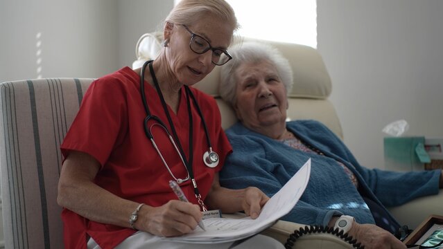 A healthcare provider in red scrubs assists an elderly woman in a cozy chair, taking notes while ensuring her well-being and comfort in a warm environment.
