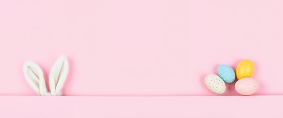 Minimalist playful scene with white fluffy bunny ears peeking from bottom against solid pink background. Pastel Easter eggs in pink, blue, yellow, speckled arranged in semi-circle below.

