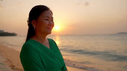 A Southeast Asian woman stands relaxed in a green dress, appreciating the beautiful sunset on a tranquil beach, surrounded by shimmering waves and gentle light