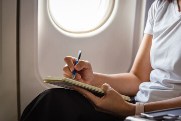 Woman hand writing on notebook in airplane with plane window background., passenger taking note in...