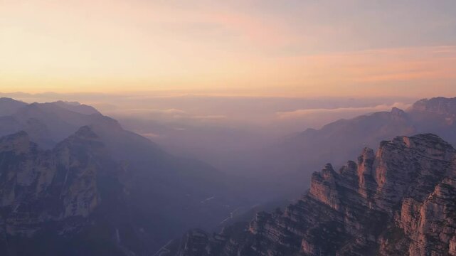 Aerial drone flying over distant mountain peaks during sunrise, casting a golden glow across the landscape and revealing vast natural beauty and majestic wilderness in the horizon
