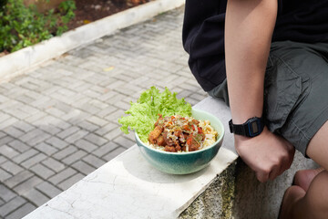 Indonesian Spicy Chicken Matah, Bowl of Mixed Salad With Crispy Toppings Placed on a Stone Ledge Beside a Person in Casual Attire Outdoors