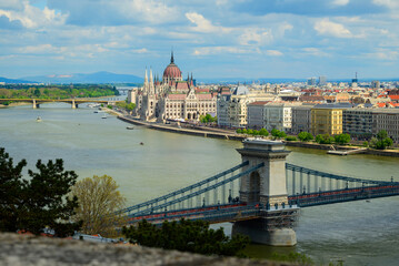 A panoramic view captures Budapest's iconic Parliament building along the Danube River, with the Chain Bridge elegantly spanning the water under a bright sky.