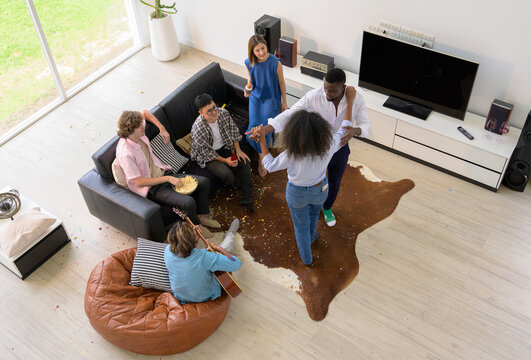 Lively group of diverse friends gathering in modern living room at home. Multiethnic celebrating with confetti scattered around while one person plays guitar and another enjoy dancing and eating