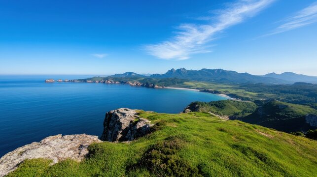 A breathtaking view from elevated land reveals a vast blue ocean meeting a rocky coastline. Lush greenery blankets the area, showcasing distant mountains under a clear sky