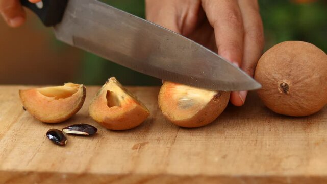 Person cutting fresh sapodilla fruit with knife on wooden board. The juicy tropical fruit is being prepared, with visible seeds and natural texture. 