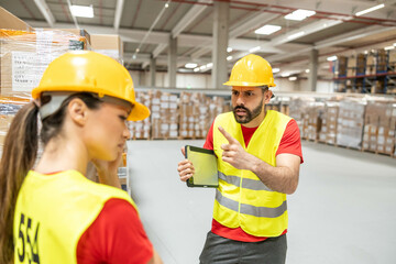 Male warehouse supervisor pointing and reprimanding a female worker near stacked pallets.