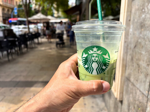 Alicante, Spain - May 16, 2025: Hand holding Starbucks iced green tea beverage in a vibrant outdoor cafe setting, showcasing refreshing drink and lively atmosphere