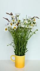 On the white table in a yellow mug there is a magnificent bouquet of daisies and other wild flowers.