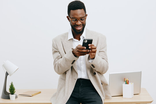 Confident smiling man with dark skin and glasses using smartphone in modern office, casual business attire, illuminated workspace, engaging with technology, professional atmosphere, lifestyle concept.