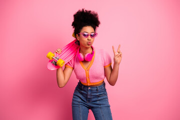 Lovely girl with afro hairstyle holding skateboard against pink background making peace gesture