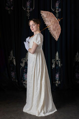 A Regency woman wearing an embroidered dress and holding a parasol, standing in front of velvet curtains