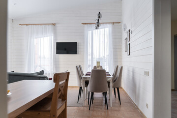design of kitchen combined with a living room in a country house. Large sofa with decorative cushions, white wooden walls and ceiling. built-in kitchen made of white wood, large windows, dining table.