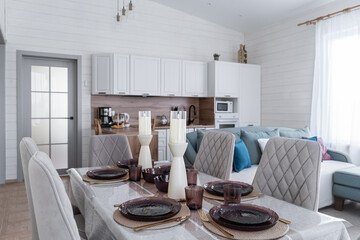 large dining table and upholstered grey chairs with backs. table is covered with linen tablecloth. dishes, glasses, candles on table. Dining room in a wooden country house. The interior of the house.